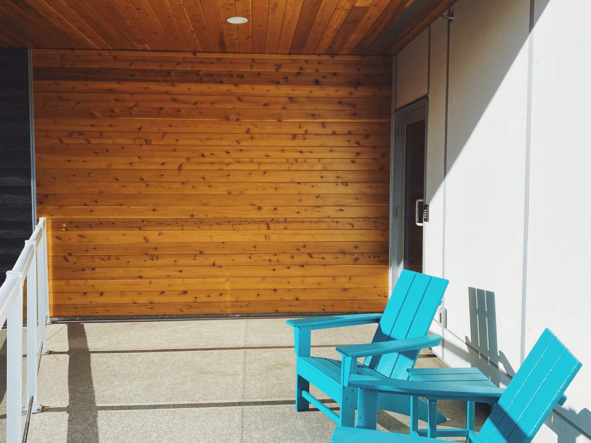 A cozy outdoor balcony area with bright turquoise wooden chairs against a warm wood-panelled wall.