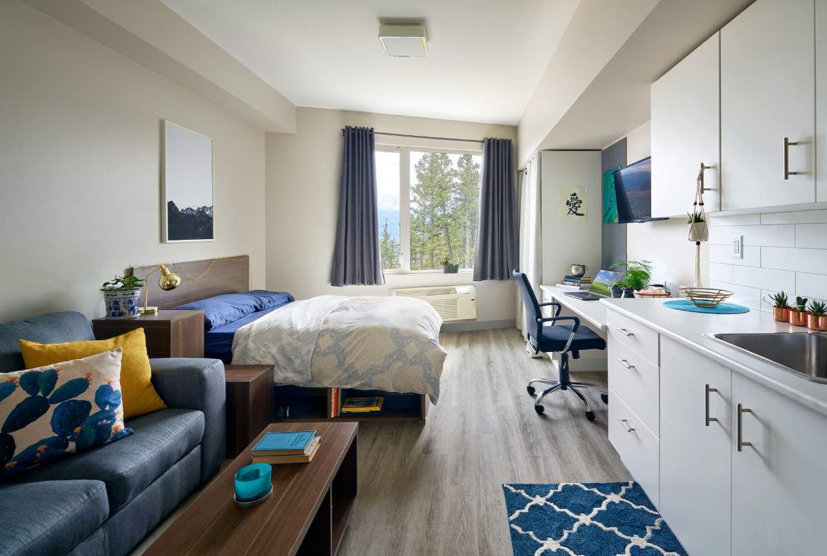 A bright and modern studio apartment featuring a bed near a large window, a sofa, and a study area with a desk and chair.