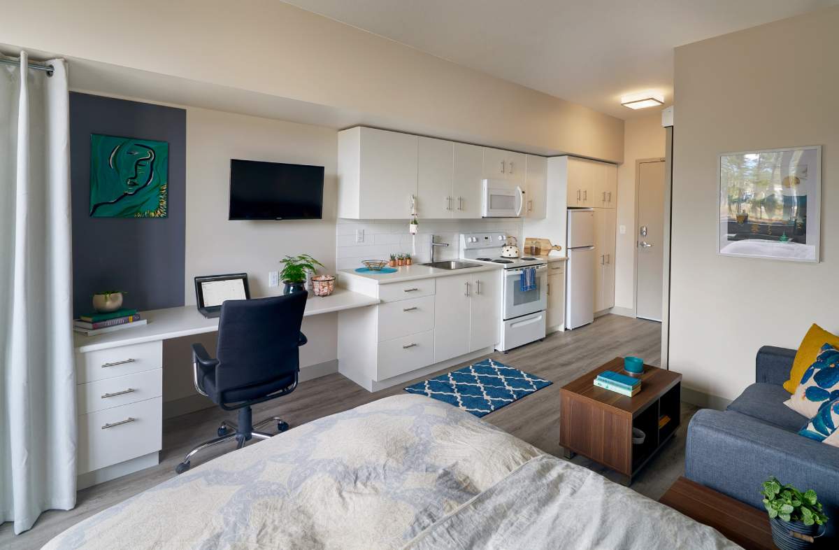 A studio apartment with a well-organized layout, featuring a fully equipped kitchenette, a study desk, and wall-mounted TV.